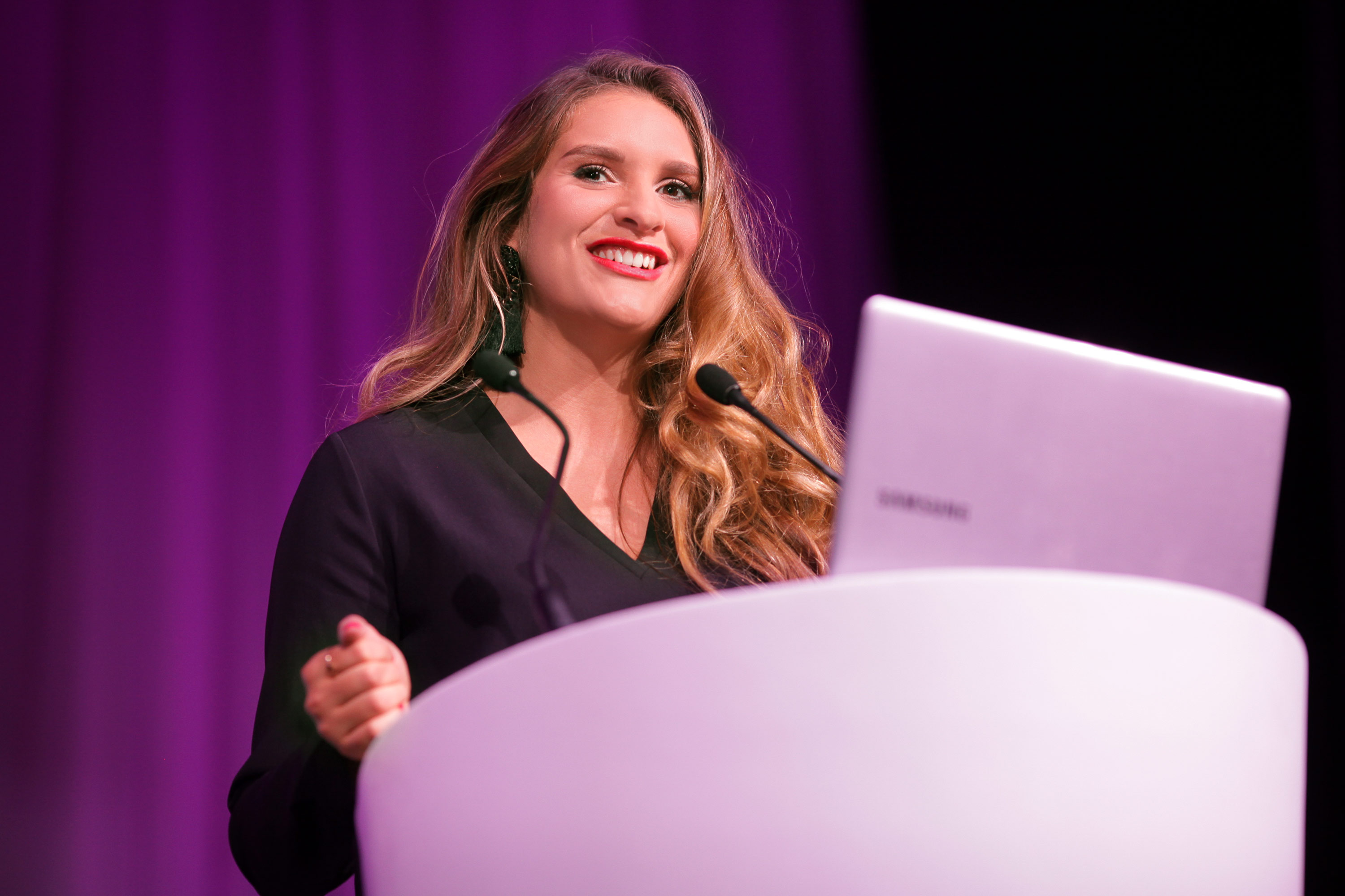Woman standing at a podium, presenting at a conference with a purple curtain backdrop