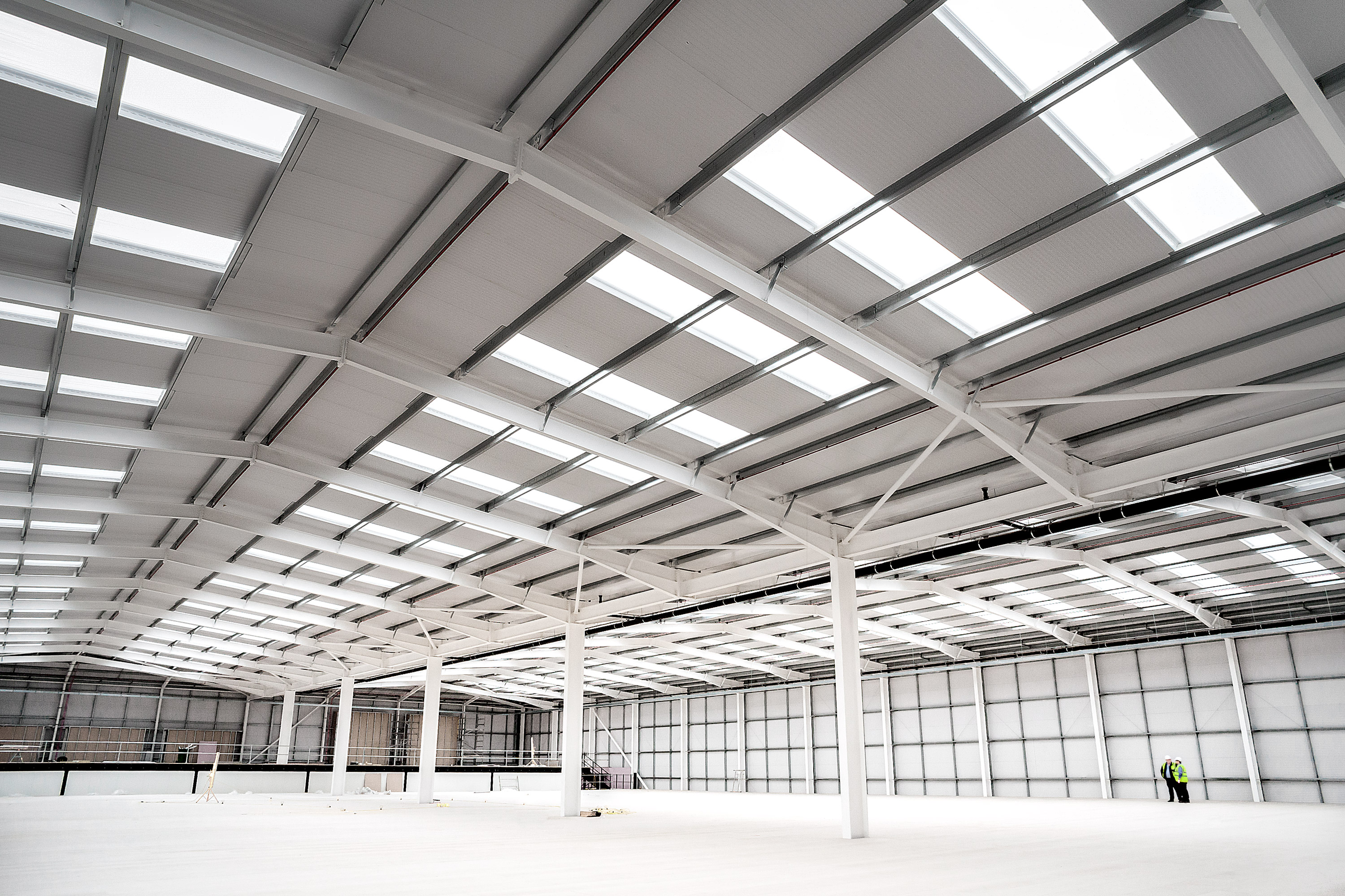 A vast, bright white empty commercial unit with workers in hard hats surveying the space