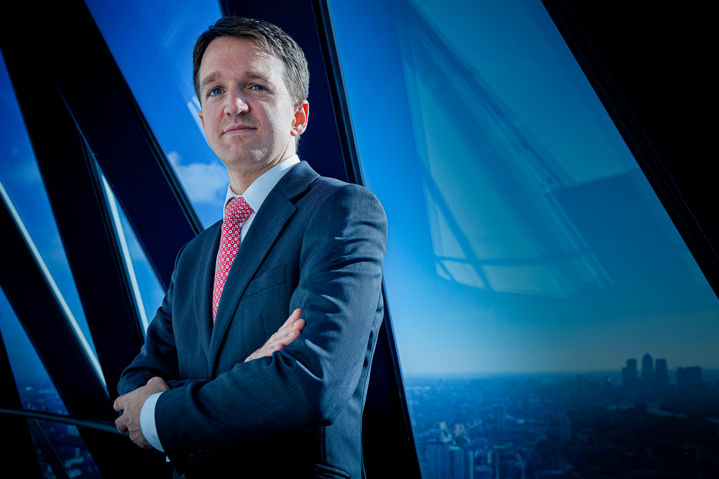A headshot portrait of a suited man in front of blue windows looking out over a cityscape from a skyscraper